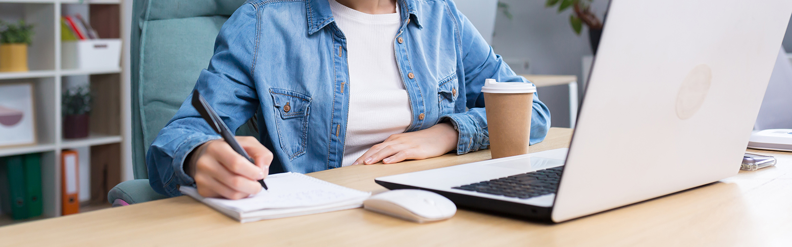 Woman sitting at a desk writing in a notebook with a laptop in front of her.